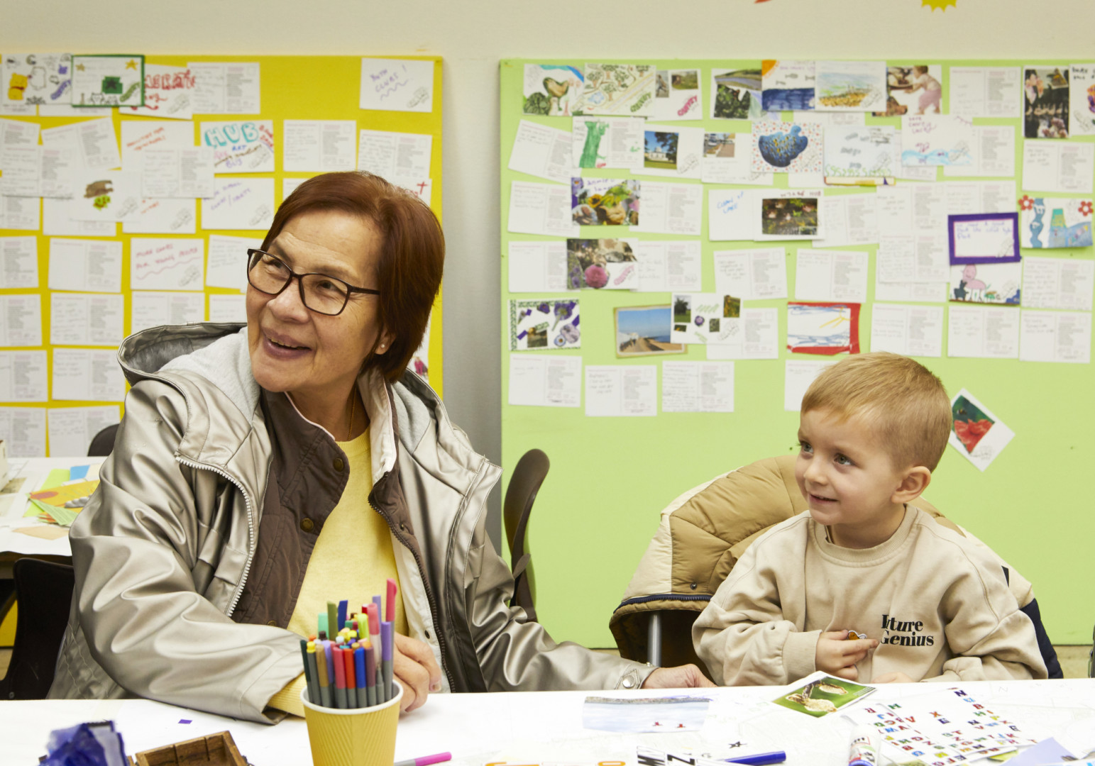 A woman and child at a table with pens and paper smiling with postcards on display in the background